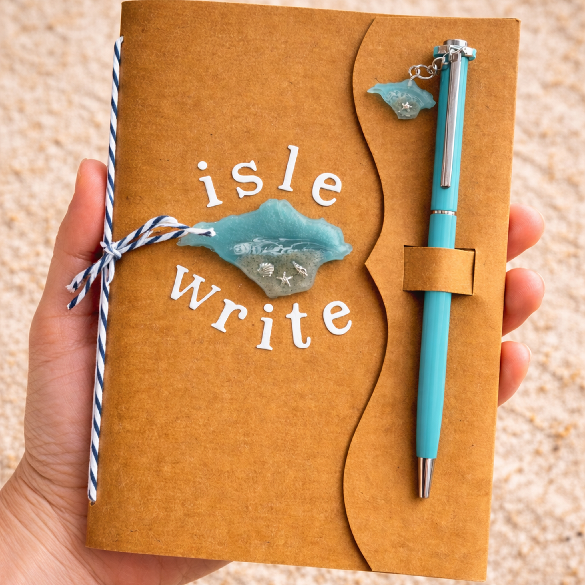Hand holding an Isle of Wight themed notebook with turquoise resin island design, beach sand detail and matching pen with charm