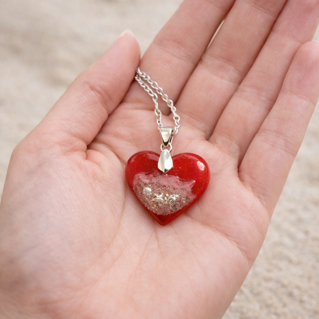Handmade turquoise resin heart pendant with real beach sand and silver shell details resting in a hand on the beach.