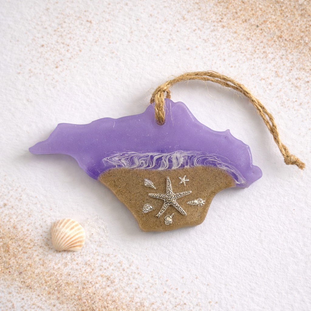 Handmade Isle of Wight shaped resin ornament with real beach sand, silver starfish and seashell details on a white background with scattered sand and a small shell.
