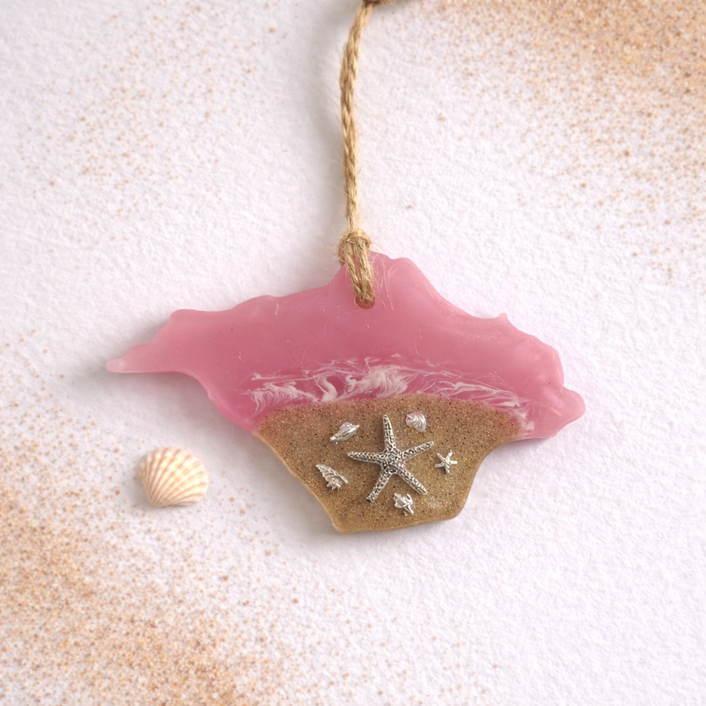 Handmade Isle of Wight shaped resin ornament with real beach sand, silver starfish and seashell details on a white background with scattered sand and a small shell.