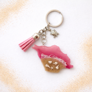 Isle of Wight pink resin keyring with real sand, silver shell charms and tassel on a white background with scattered sand