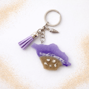 Isle of Wight purple resin keyring with real sand, silver shell charms and tassel on a white background with scattered sand